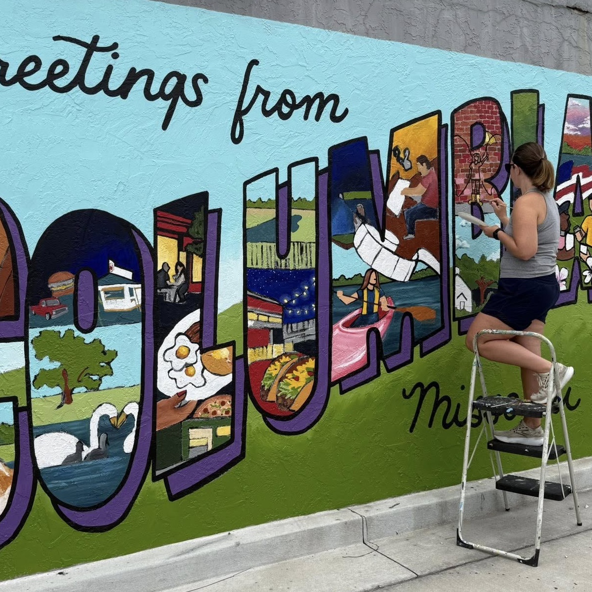 A woman standing on a ladder painting a Columbia mural.